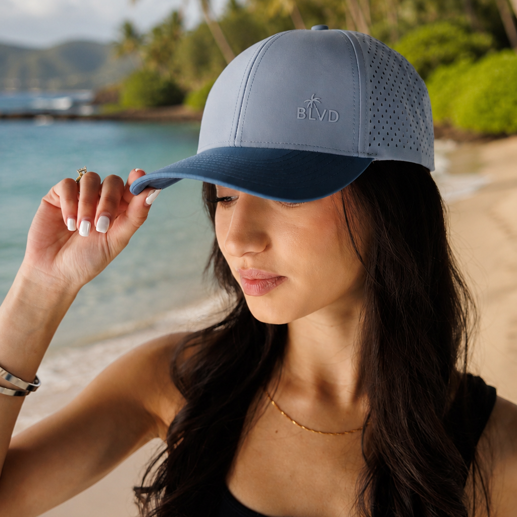 Woman holding a gray cap with 'ALOHA' text on a beach with palm trees and water in the background